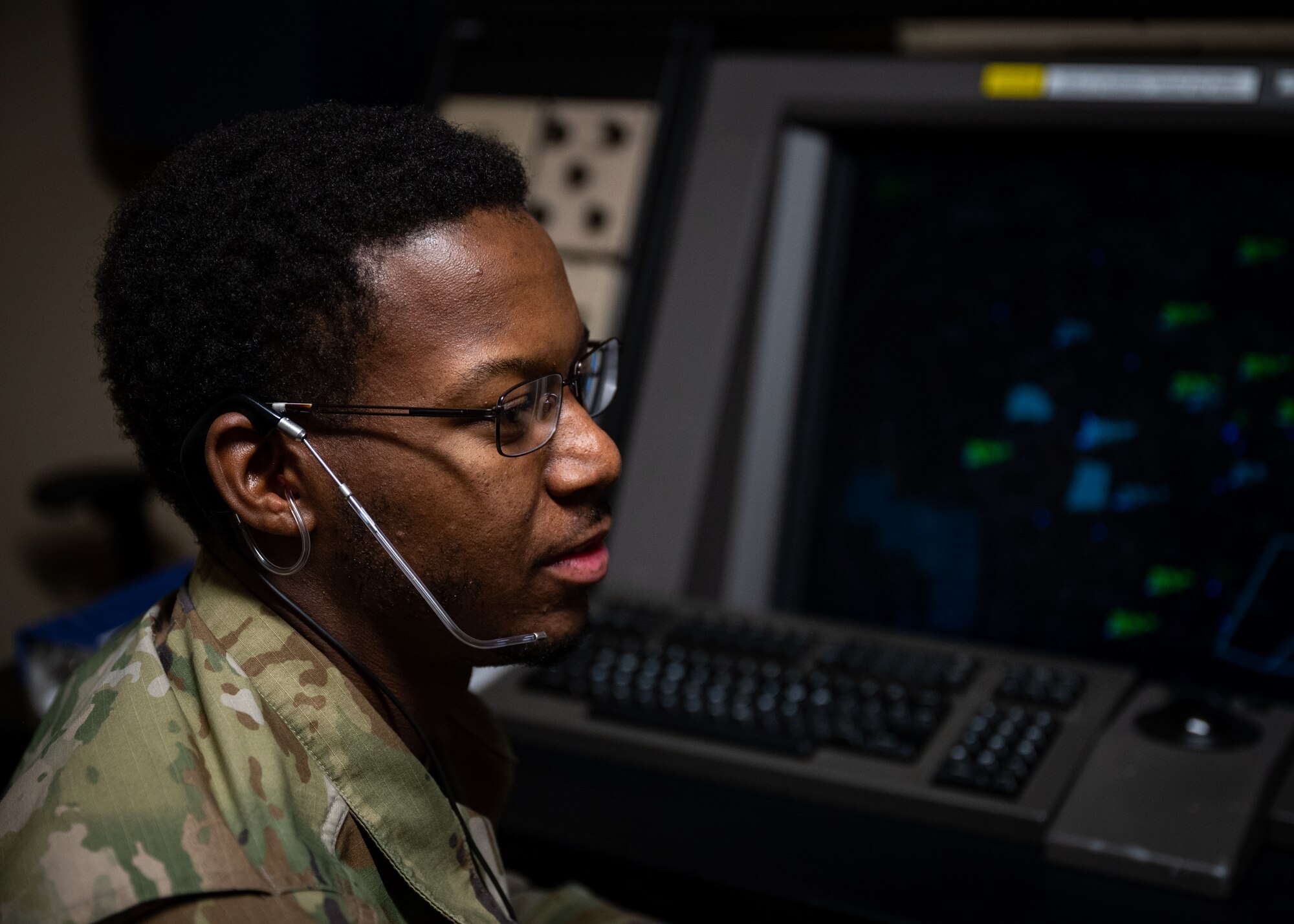 Male Airman wearing a headset looks to the right in front of a dark screen with blue and green aircraft markers