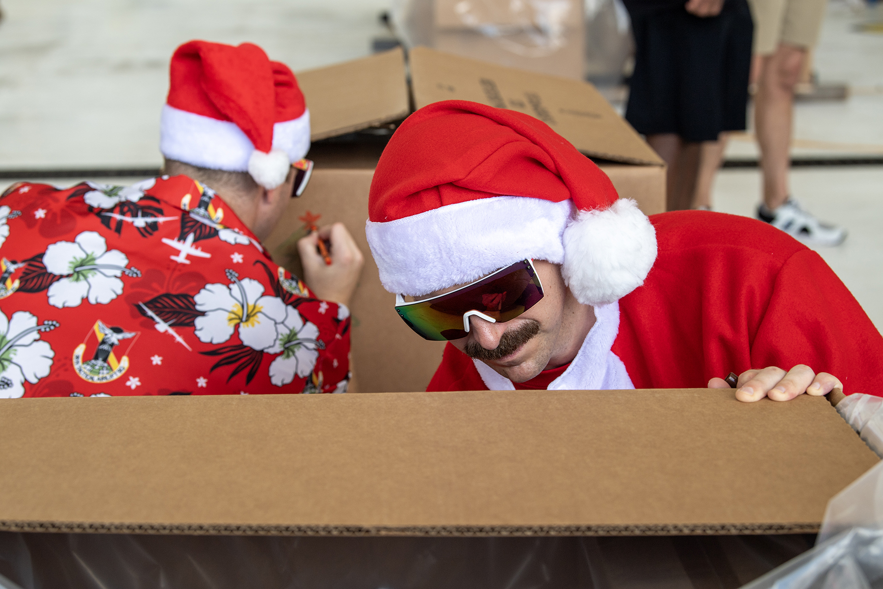 Two service members wearing Santa hats crouch while coloring on cardboard boxes.