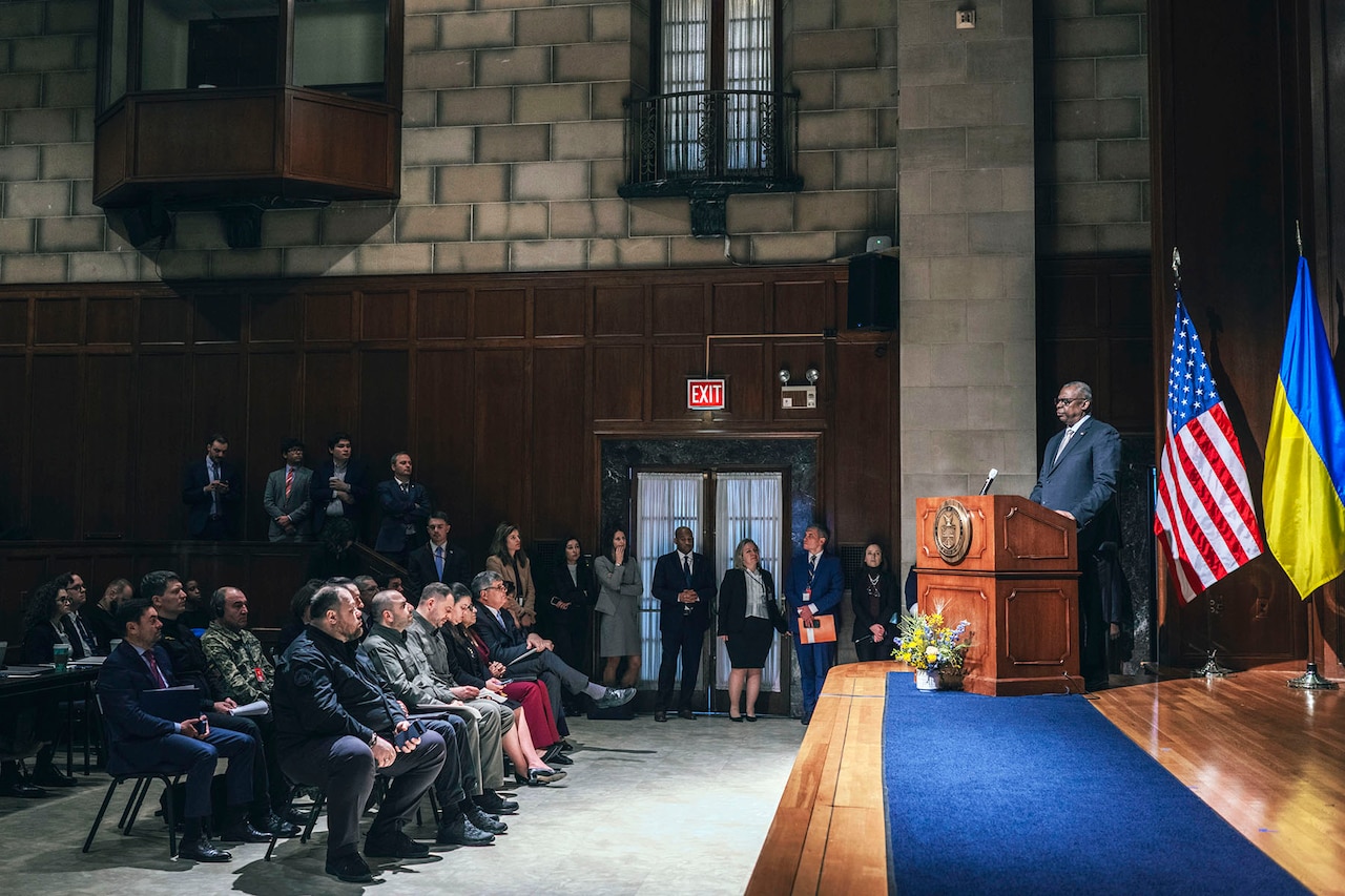 A man in a suit stands at a lectern with U.S. and Ukrainian flags in the background.