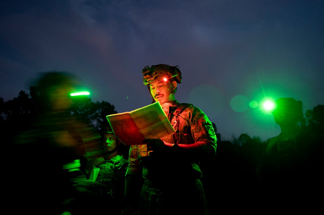 A service member, illuminated by green lights, reads a map at night as other service members stand nearby but are out of focus.