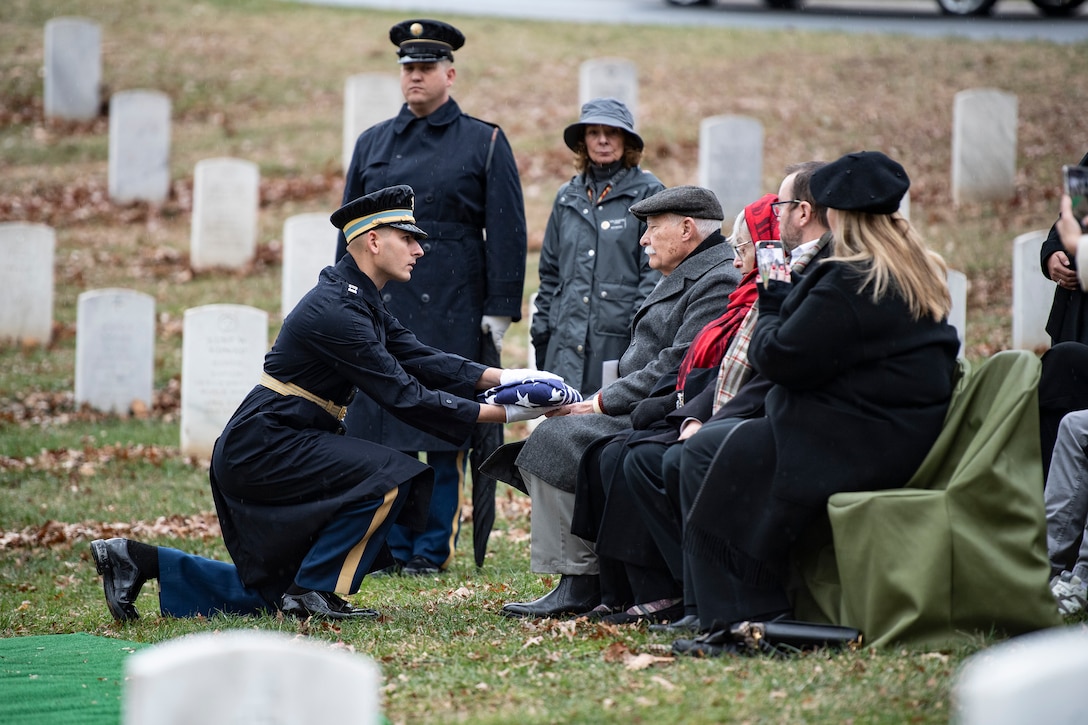 A service member kneels to present the American flag to a seated person in a cemetery as others are seated nearby.