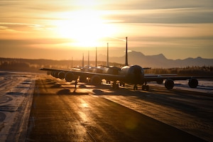 Military aircraft form a line on the tarmac with low sunlight in the background.