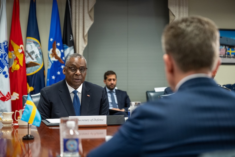 A man in a suit is seated at a table speaking with another seated man.