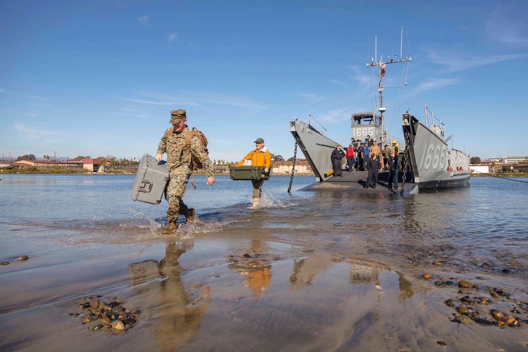 Service members walk through waters while carrying bins as a vessel moors in the background.