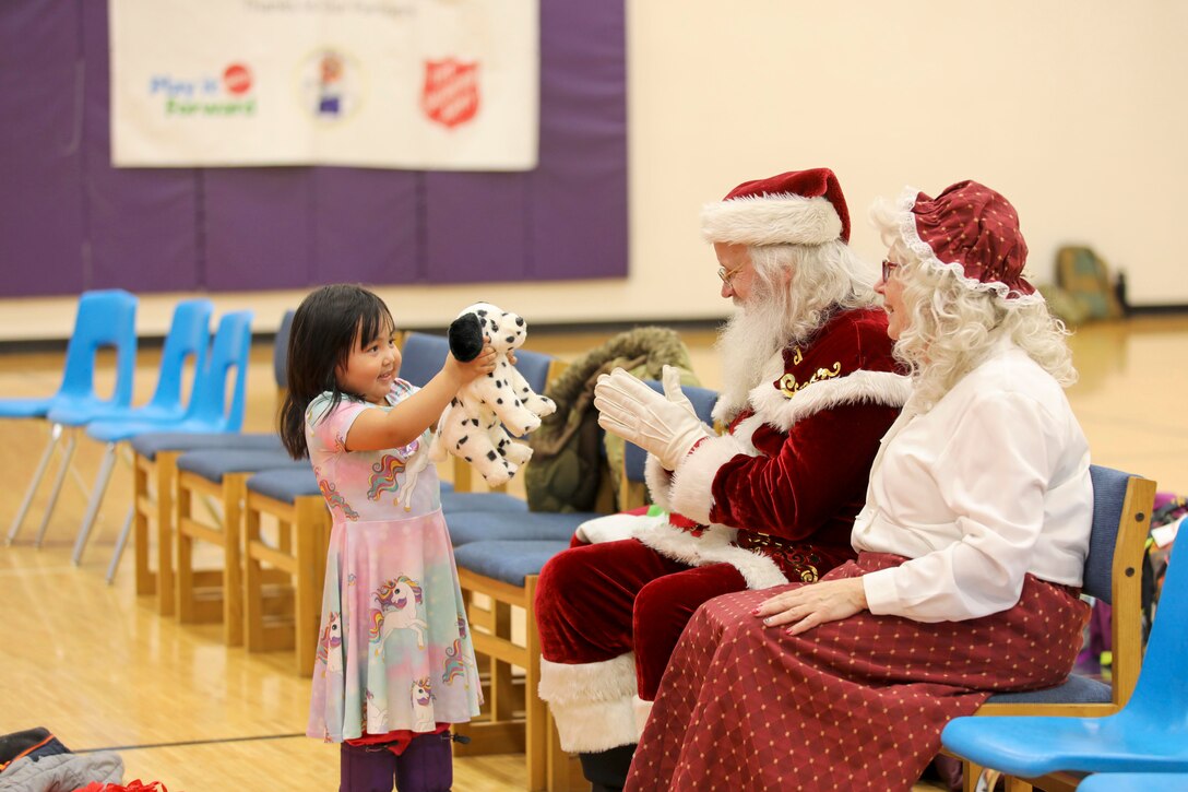 A child shows a stuffed animal to Santa and Mrs. Claus while in a gymnasium.