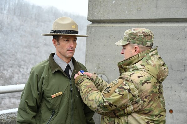 A U.S. Army Officer pins a blue medal on a U.S. Army Corps of Engineers Ranger atop a concrete dam with snow falling.