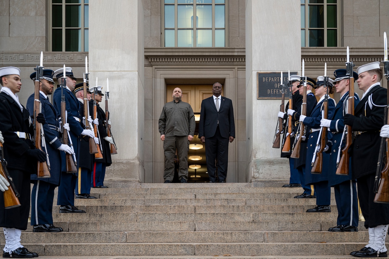 Two men stand at the top of stairs as an honor guard greets them.