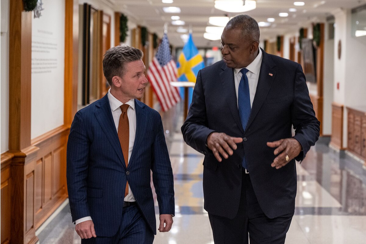 Two men wearing suits walk down a hallway.