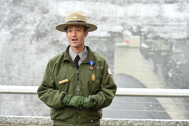 A U.S. Army Corps of Engineers Ranger stands atop a concrete dam with snow falling.