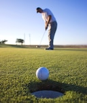 close up of golf ball sitting on the edge of a hole with golfer in the background