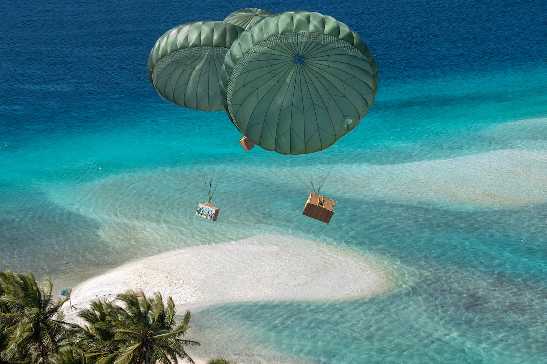 Boxes attached to parachutes descend toward a beach during daylight.