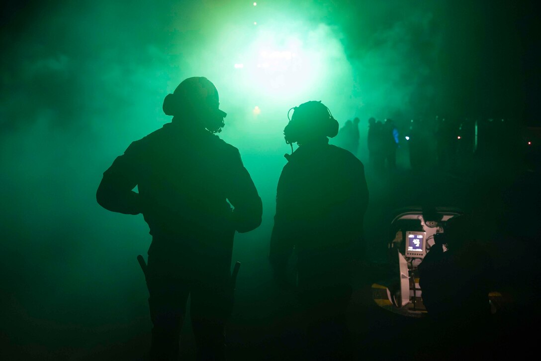 Two sailors shown in silhouette are illuminated by green light as others stand in front of them.