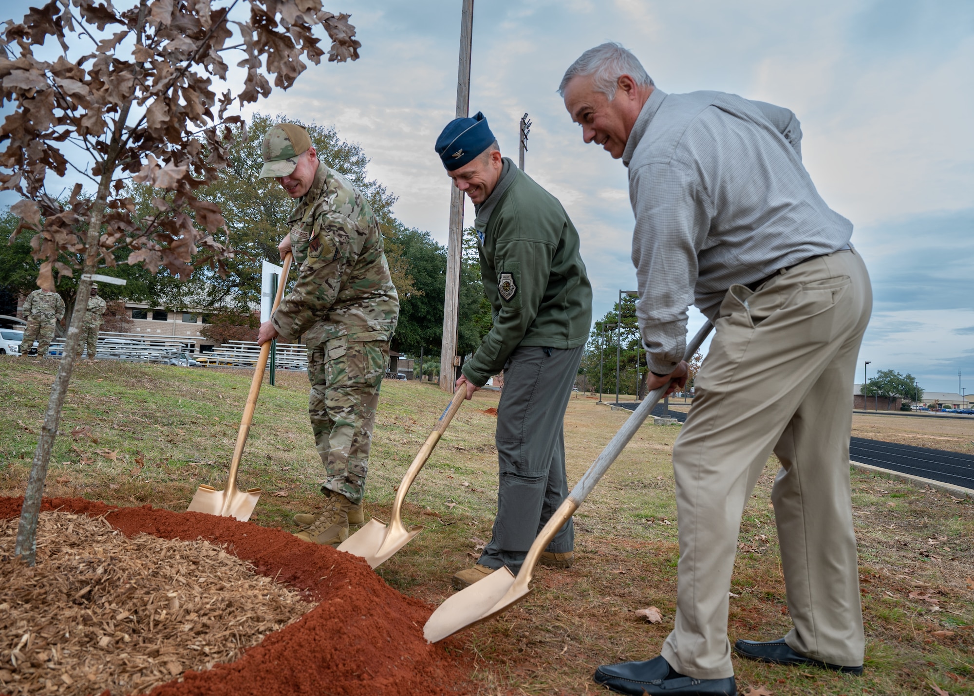 Three men lean down to dump dirt from their shovels around the base of a tree.