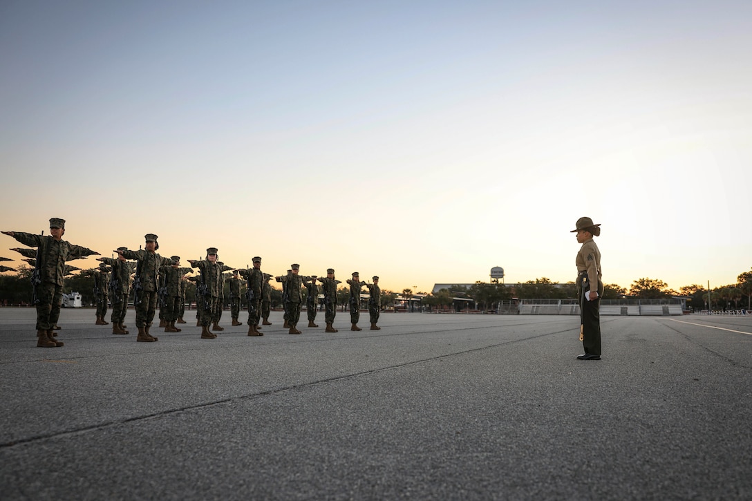 A drill instructor watches as dozens of recruits stand in formation holding up their arms in a parking lot under a sunlit sky.