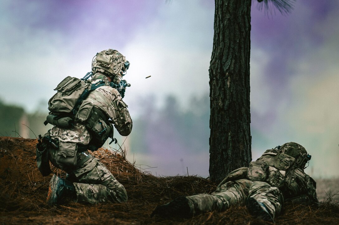 A kneeling soldier fires a weapon into clouds of yellow and purple smoke as a fellow soldier laying on the ground watches.