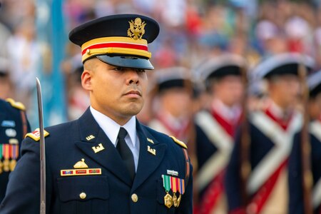 An Army soldier dressed in dark blue dress uniform is standing at attention with a sword in his right hand that is resting against his shoulder. There are others in the distance.