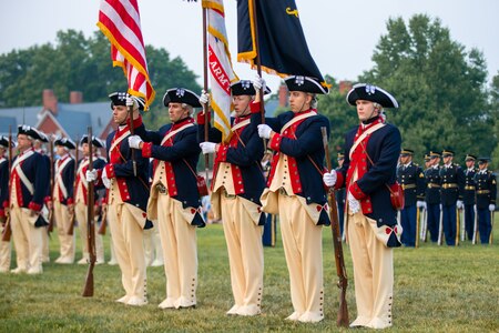 A colors team dressed in dark blue jackets and white pants that are from the Revolutionary War period are holding three flags and the members on the ends are holding muskets. There are other people in the distance behind them.