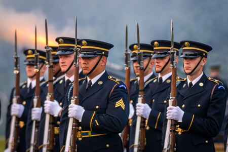 Seven Army Soldiers are in dark dress uniforms while holding files out in front of their bodies with bayonets affixed to the end of the rifles.