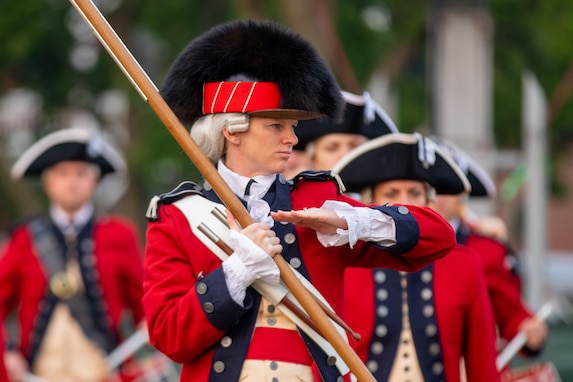Someone dressed in a red, Revolutionary War-era uniform with a large, black, furry hat is holding a long pole while making a flat-handed gesture with the other hand. Several other members of the group are behind the leader of the group.
