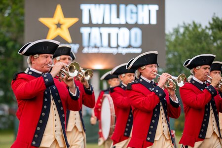 Several musicians wearing red Revolutionary War-era uniforms with dark blue tri-cornered hats are playing bugles and drums in front of a sign that reads Twilight Tattoo.