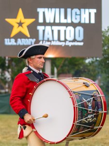 A drummer who is holding a bass drum is dressed in a Revolutionary War-era uniform that is red and includes a dark blue tri-cornered hat. In the distance behind him is a sign that reads U.S. Army Twilight Tattoo.