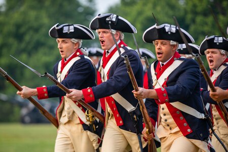 Several Soldiers wearing Revolutionary War-ear dark blue uniforms are running with rifles in hand. The rifles have bayonets affixed to them.