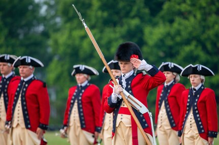 Several people are dressed in red Revolutionary War-era uniforms. The one in the front of the group is wearing a large, furry hat while holding a long wooden staff with a silver tip in one hand and saluting with the other hand.