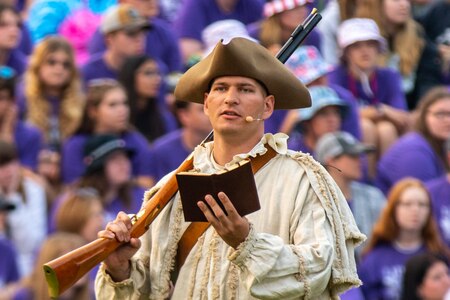 Someone wearing a Revolutionary War-era three-cornered hat is holding a small book in one hand and a rifle is slung over his shoulder in the other hand. There are lots of people in normal attire in the distance behind him.