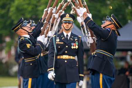 An Army Soldier is carrying a sword while walking underneath an arch of extended rifles that other Soldiers are putting into the air above him. They are all wearing dark blue dress uniforms.