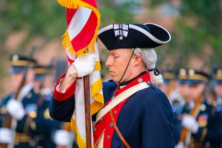 An Army soldier is holding the US flag while wearing a Revolutionary war-era blue-colored uniform and a three-cornered hat. Other Soldiers are seen in the distance.