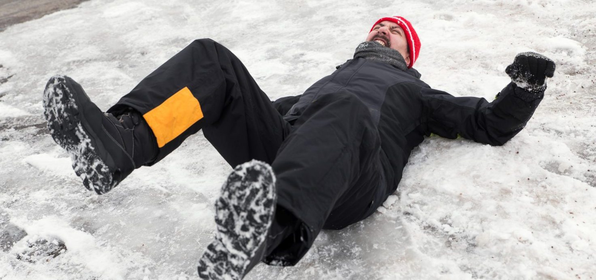 A man, on his back with legs in the air appears to slip on ice and snow