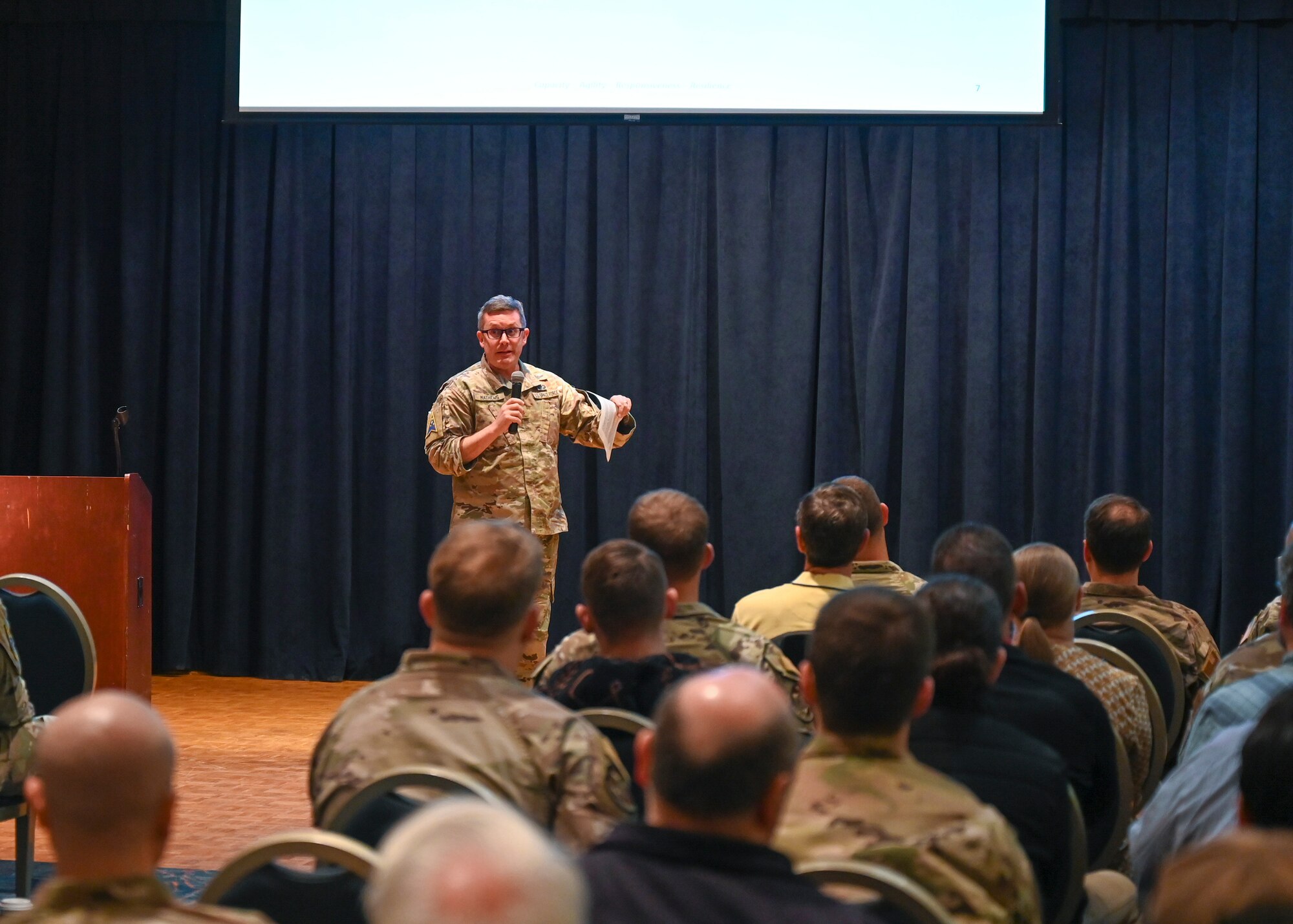 Major Mathews speaks in front of a seated crowed.