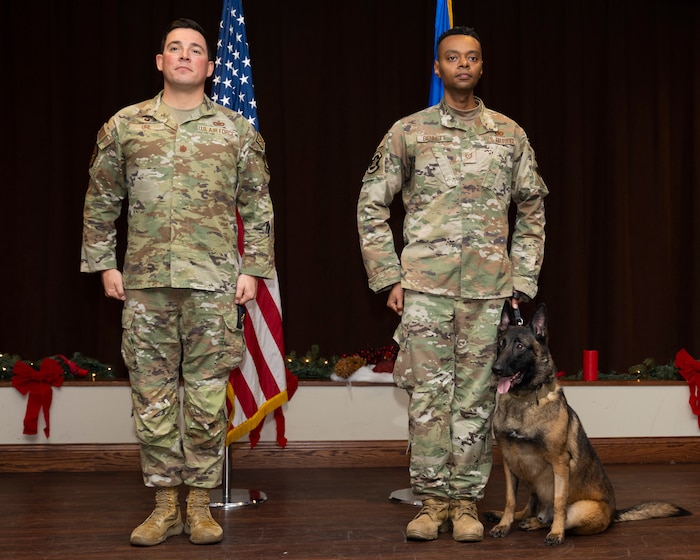 Two men in uniform stand at attention. Next to the one on the left is a dog. The American and Air Force flags are in the background.