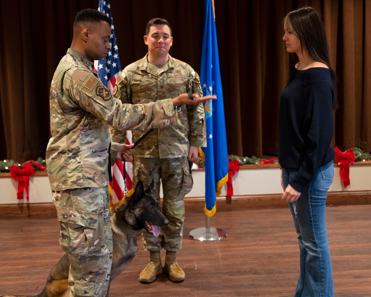 A man in uniform, with a dog next to him, holds the leash in an extended hand. A woman, in civilian clothes, waits to take the leash. Another man in uniform looks on. The American and Air Force Flags are in the background.