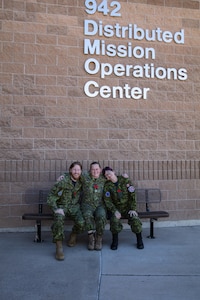 uniformed coalition military members stand sit on a bench in front of brick building with lettering “942 Distributed Mission Operations Center”