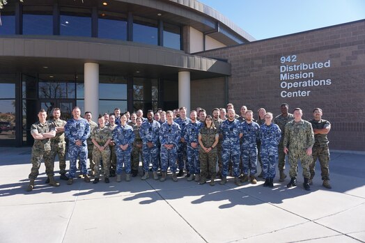  a large group of uniformed U.S., U.K. and Australian military members stand in front of brick building with lettering “942 Distributed Mission Operations Center”