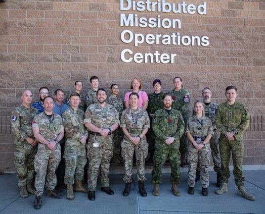  uniformed U.S., U.K. Australian and Canadian military members stand in front of brick building with lettering “942 Distributed Mission Operations Center”