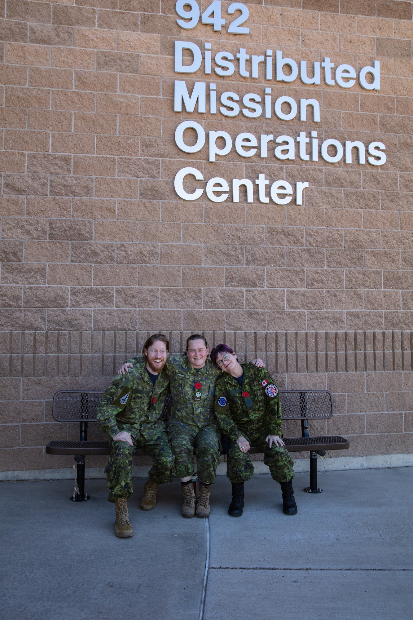 uniformed coalition military members stand sit on a bench in front of brick building with lettering “942 Distributed Mission Operations Center”