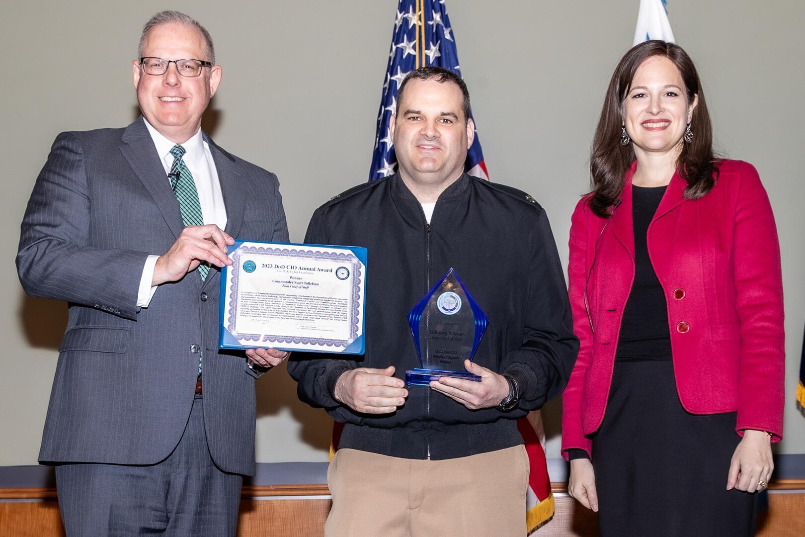 A man dressed in a military uniform stands on a stage. He is flanked by a man holding a certificate and a woman.