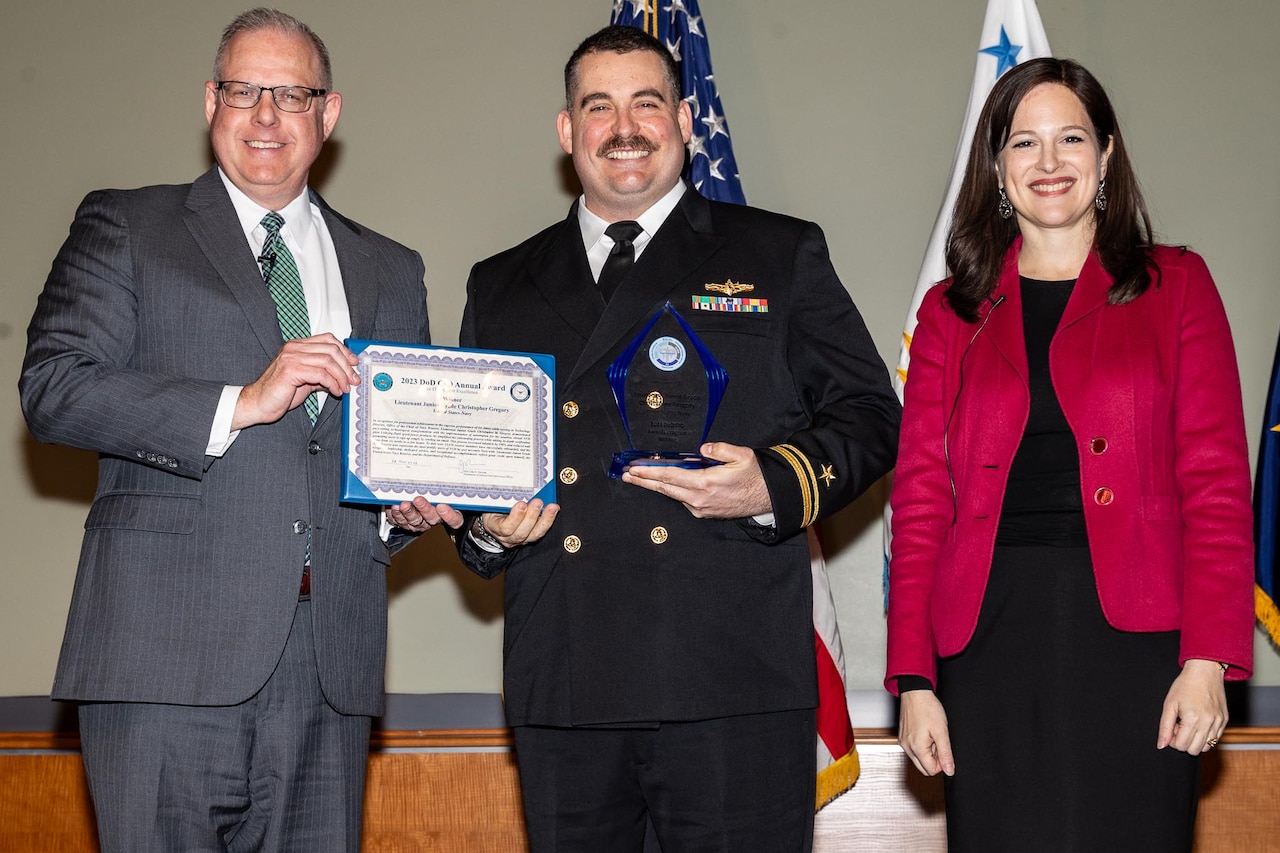 A man wearing a military uniform and holding an award is flanked by a man holding a certificate and a woman.