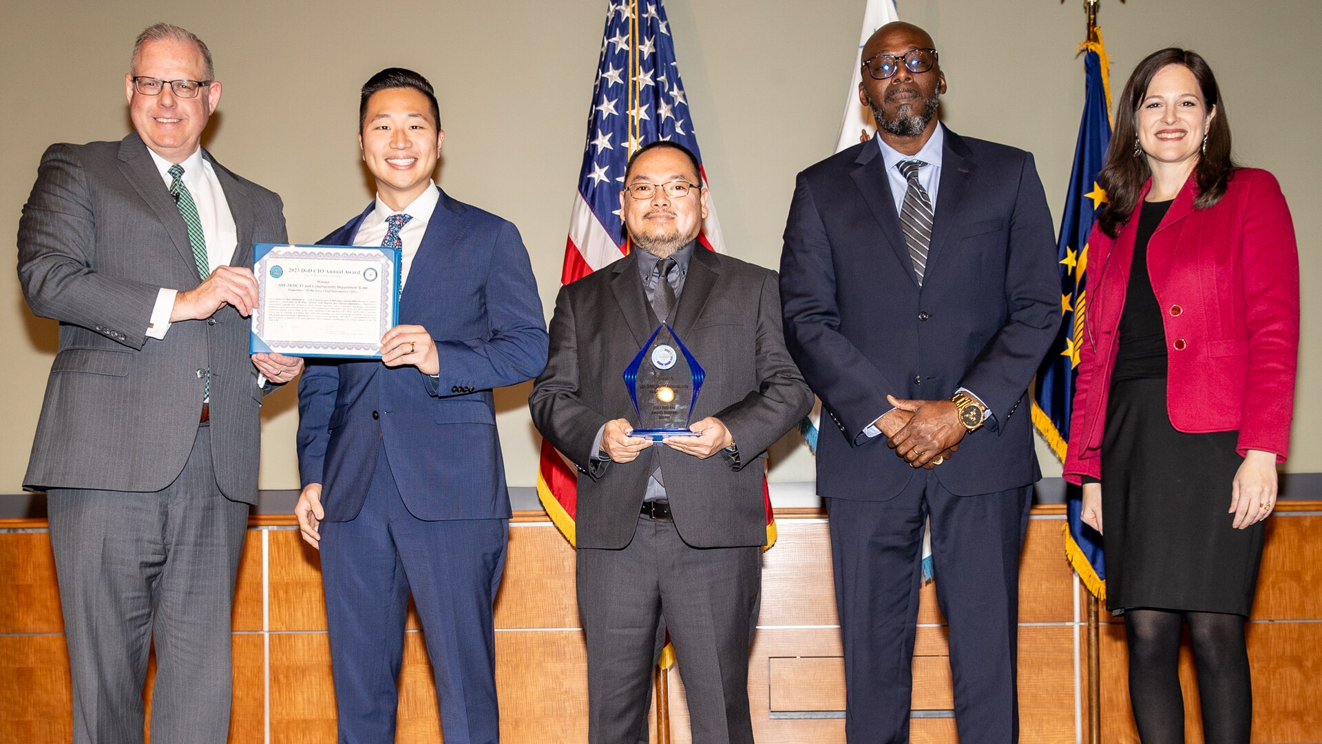 Five people stand together on a stage. One man holds a certificate. Behind the group are several flags.