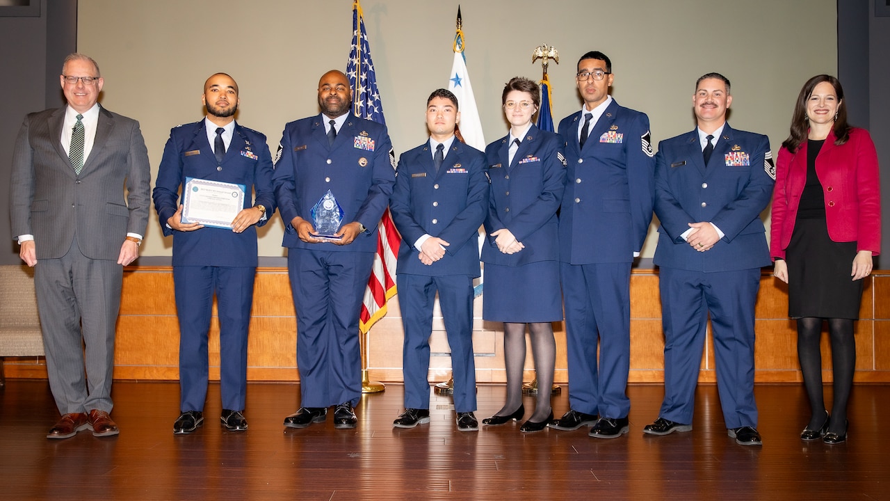 Eight people stand together on a stage. Six people are in military uniforms; the others are in civilian clothing. One man holds a certificate. Behind the group are several flags.