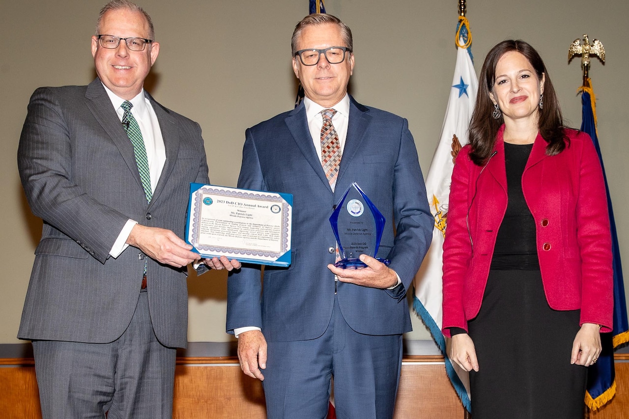 A man holding an award is flanked by a man holding a certificate and a woman.