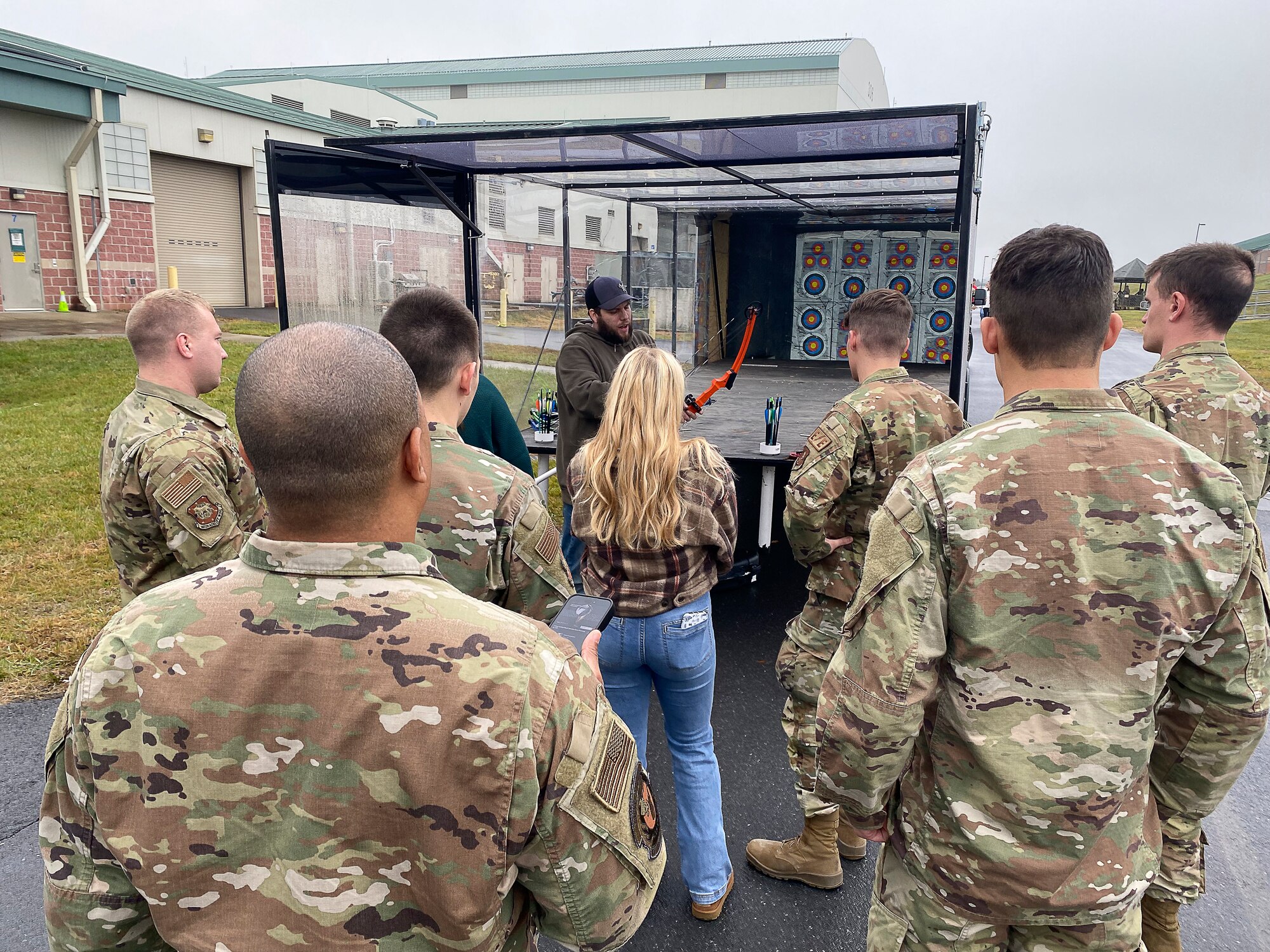 Airmen and women listen to a man give instrcutions on how to use a mobile archery range.