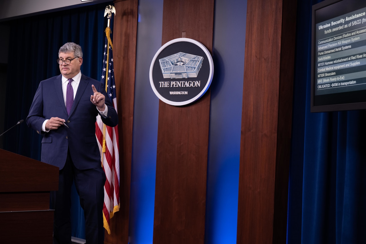 A person in a business suit gestures with their hand while standing at a lectern.