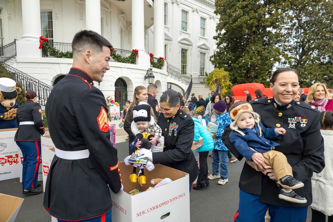 Marines carrying toddlers look inside boxes full of toys as kids in the background carry presents while walking around outside of the White House.