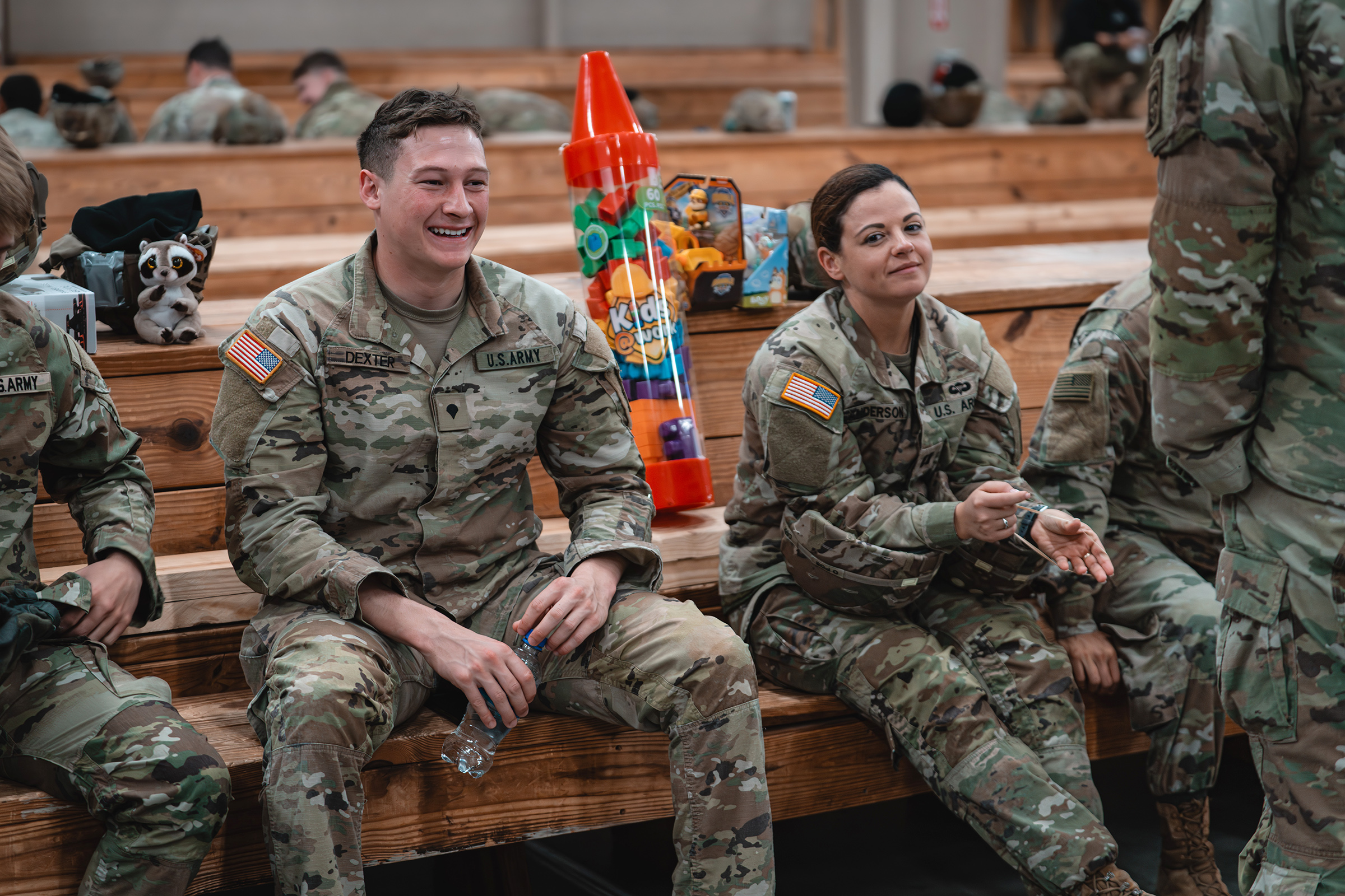 Soldiers sit on benches in a large room with toys on a table behind them. 