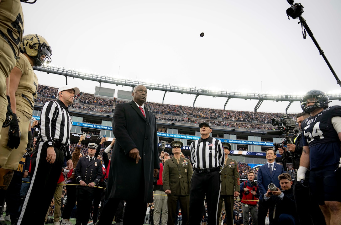 The secretary of defense tosses a coin surrounded by football players and referees.