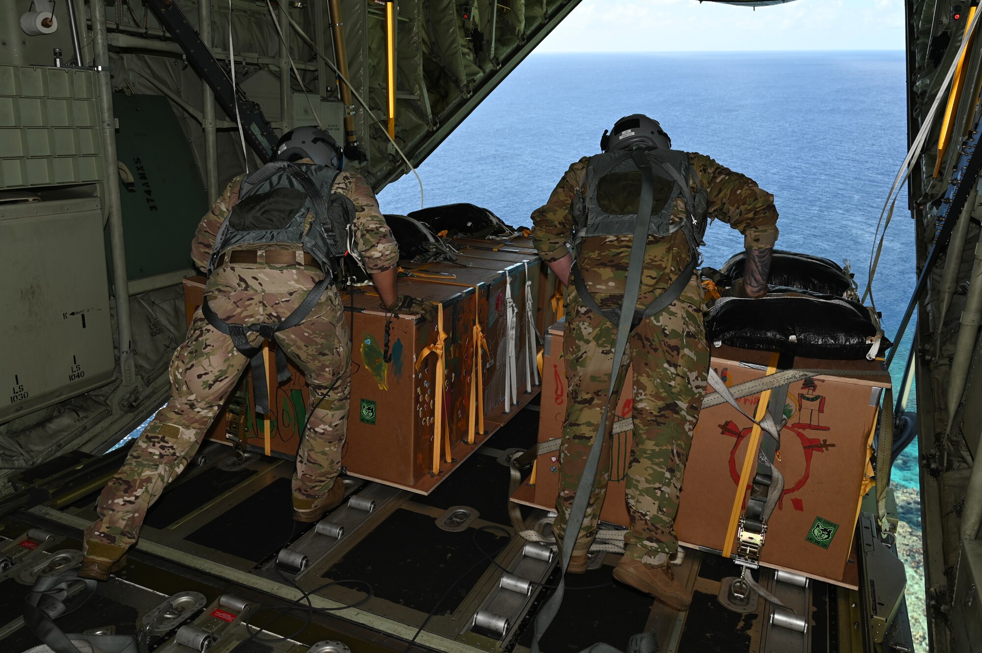 Two Airmen physically push bundles of humanitarian aid off an open C-130J Super Hercules ramp.