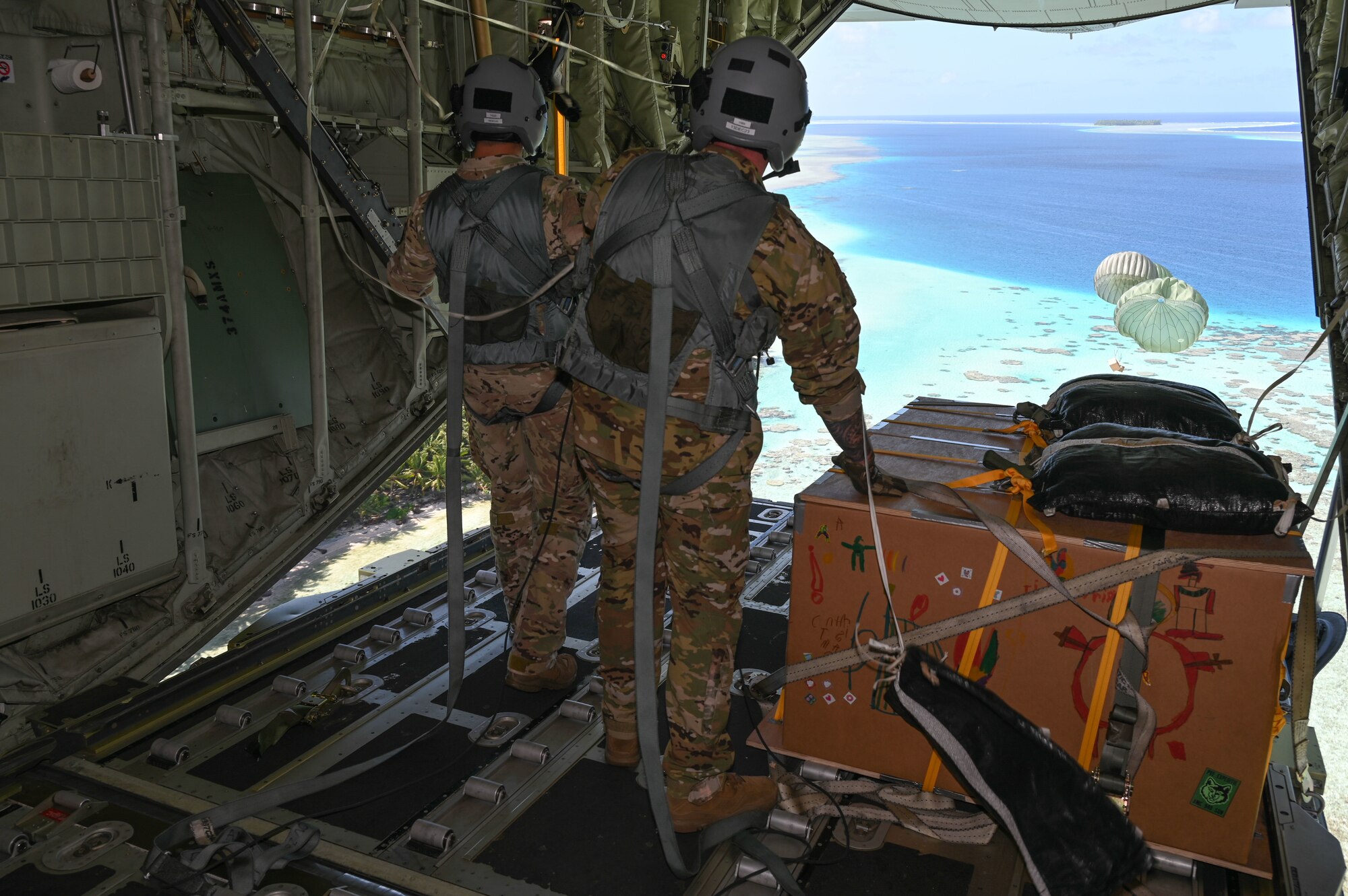 Two Airmen are on standing on an open ramp of a C-130J Hercules and watching bundles of humanitarian aid drop (with parachutes) to the island of Eauripik.